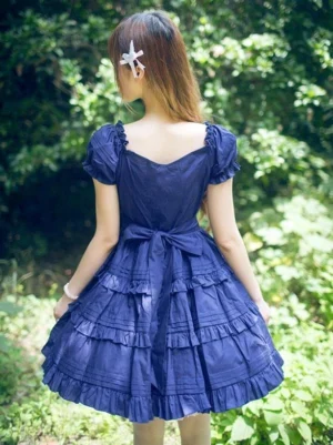 Back view of navy lolita dress showing square back neckline, waist bow detail, and multi-tiered ruffled hem on model in garden background