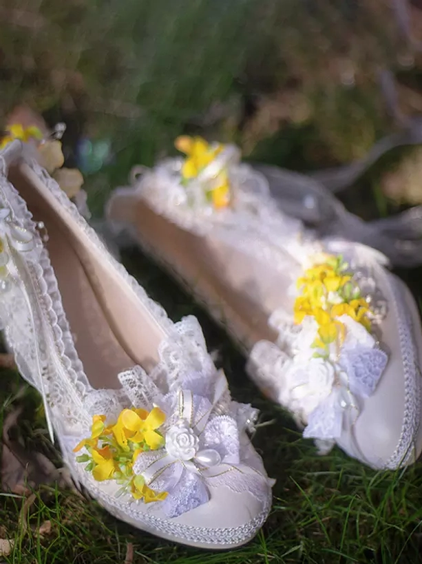 Pair of Lolita pumps with yellow flower decorations and lace bow trims on light neutral base photographed from oblique overhead angle on outdoor grassy lawn setting