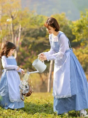 Parent-child matching blue gingham Lolita one-piece dresses with white lace-trimmed aprons and bonnets, adult woman and little girl wearing coordinated outfits outdoors in grassland setting with sheer white long inner sleeves visible