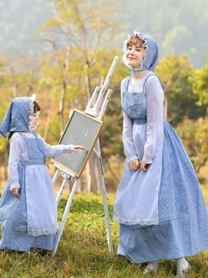 Matching maid-style Lolita dresses in blue gingham with white lace-trimmed aprons and coordinating bonnets, parent and child models posed beside outdoor painting easel showing side-front angle with sheer white inner blouses