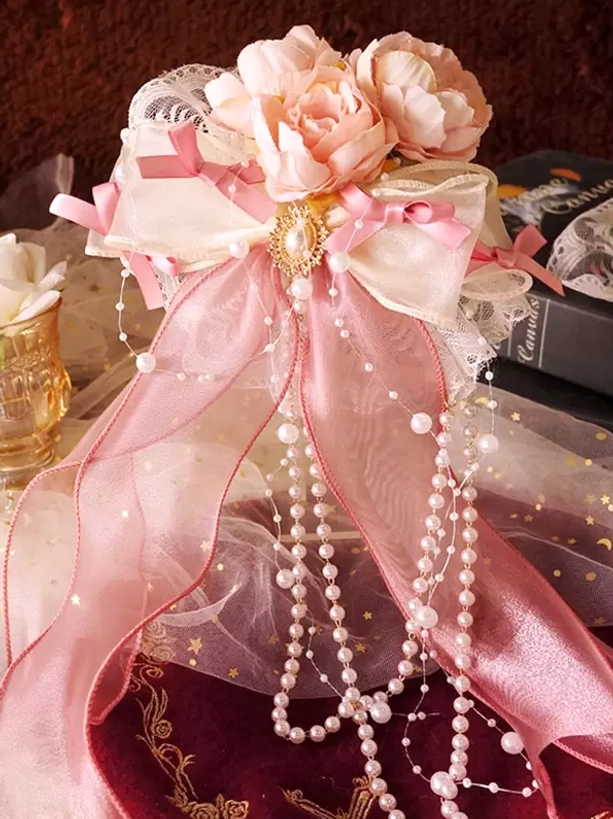 Close-up front view of peach pink lolita hair accessory featuring beige-pink ribbons, artificial flowers, golden brooch accent and lustrous pearl pendants, styled on velvet with vintage book prop for elegant presentation