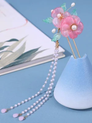 Pink floral tassel hairpin with gold centers and pearl accents displayed on decorative blue vase and notebook, overhead oblique view showing resin flower details and cascading pearl tassel design
