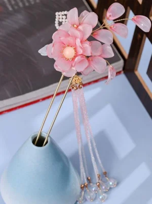 Pink floral tassel hairpin with layered resin blooms and gold metal hair fork displayed on ceramic vase with books and wooden frame background - lolita hair accessory three-quarter view