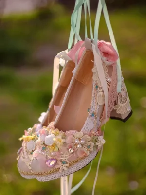 Top-down overhead hanging view of single pink lace bridal lolita heel shoe displaying decorated lace trims gemstone accents and pink fabric bow detail against blurred green outdoor background