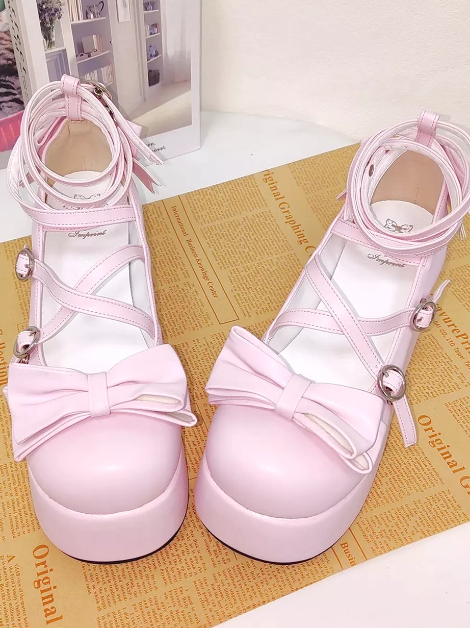 Pink Lolita platform pumps front flat lay view with newspaper print backdrop showing large front bows, rounded closed toe shape, cross ankle straps, and decorative embellishments