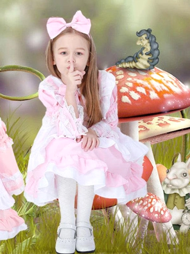 Pink maid lolita dress with puffed sleeves, ruffled lace details and large pink bow worn by child model seated in fairy tale mushroom garden aesthetic setting