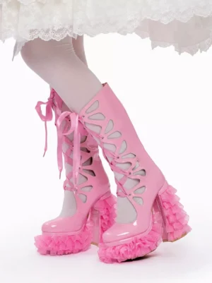 Pink patent leather sweet Lolita platform boots with criss-cross ribbon lacing shown in front three-quarter view on model wearing white lace skirt, featuring side cutouts and ruffled block heel