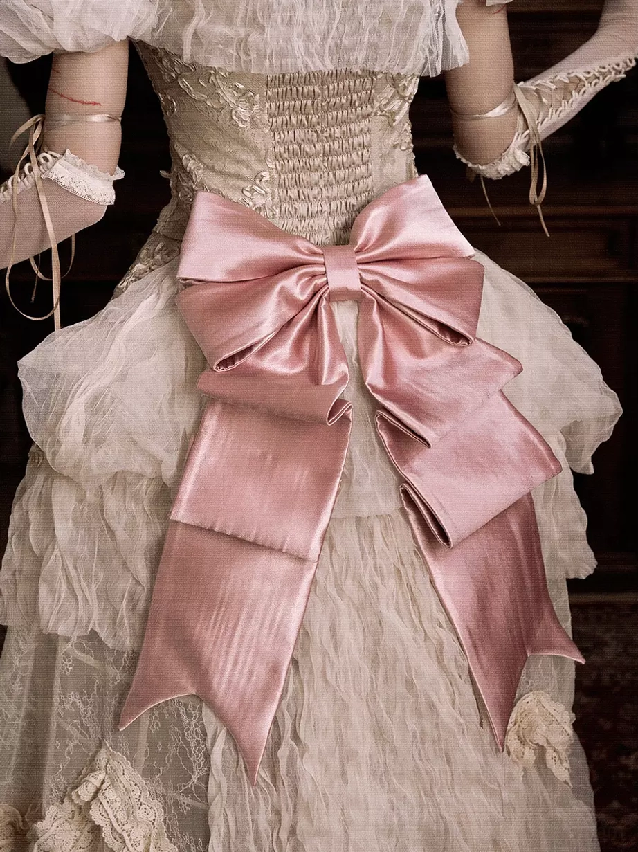 Side rear close-up detail of shiny double-layer pink satin bow on vintage wedding dress with smocked corset bodice, showing intricate bow structure and ribbon tail flow in atmospheric lighting