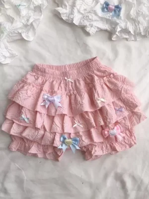 Top-down flat lay of pink ruffled lolita shorts featuring mixed pastel bow decorations and layered ruffled hem design on white fabric background