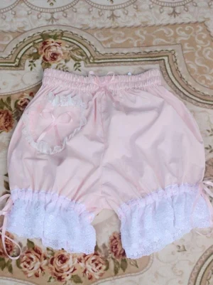 Pink lolita bloomer shorts with light purple lace ruffled hems and coordinating satin bow pocket, flat lay photography on vintage floral background, demonstrating versatile color option for sweet lolita coordinates, comfortable fit design
