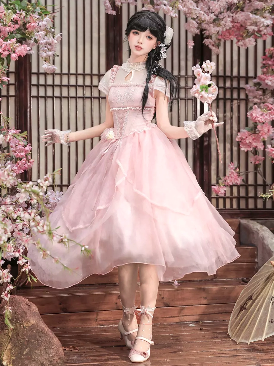 Full body front view of pink sweet lolita dress featuring short puff sleeves, lace-up corset top, and voluminous tulle skirt worn on terrace with cherry blossoms