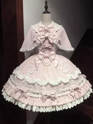 Pink sweet lolita JSK dress with detachable lace-trimmed cape, full front view on mannequin showcasing multi-layered ruffled skirt, pearl necklace trim, and large front bow