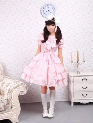 Pink sweet lolita OP dress with tiered ruffles and lace-up bodice worn by model in full front view, featuring white socks and hair accessory in vintage interior setting
