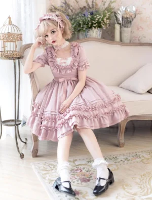 Pink sweet lolita OP dress with cascading ruffled skirt and short puffed sleeves, worn by blonde model in vintage indoor setting, front three-quarter sitting view showcasing romantic chiffon silhouette