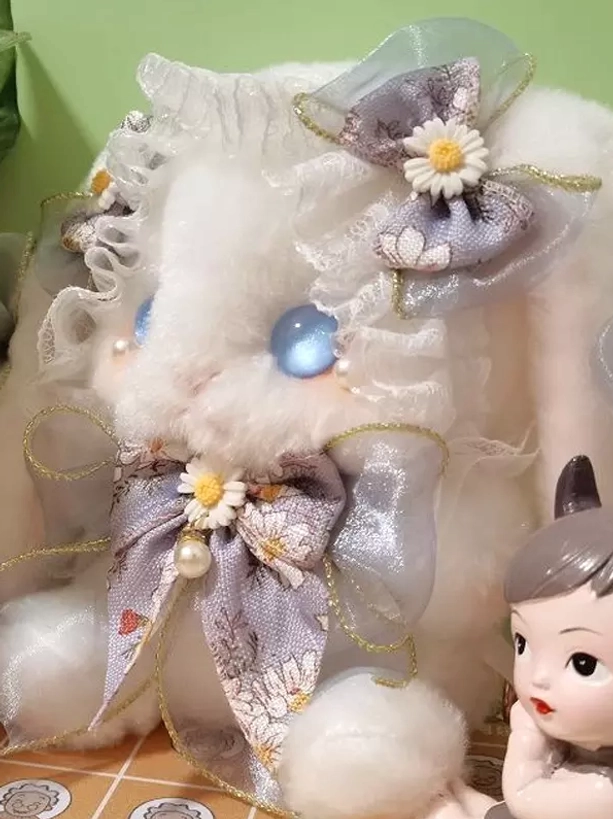 Plush bunny lolita bag with purple daisy bow, blue glass eyes, and ruffled lace trim displayed on tabletop with small doll ornament for scale