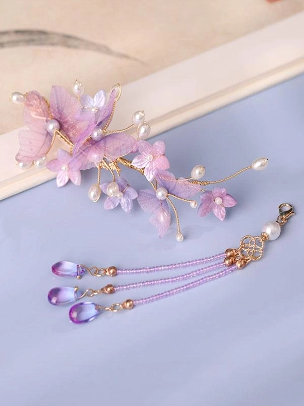 Flat lay overhead view of long tassel purple floral hair accessory with multiple sheer purple flowers gold wire branches pearl decorations and purple gradient water-drop tassel on light purple backdrop