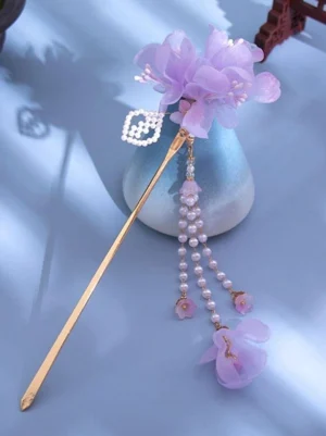 Purple flower tassel hairpin with pearl rhombus charm displayed in oblique top-down view, featuring layered chiffon purple flowers, gold hair stick, and cascading pearl tassel with flower pendants, styled against blue ceramic vase and Chinese wooden decor