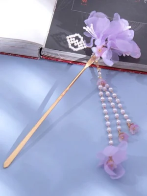 Side angle view of purple flower lolita hairpin showing detailed pearl rhombus ornament, sheer chiffon purple blooms, and flowing pearl tassel with flower charms, photographed with open book and light blue background