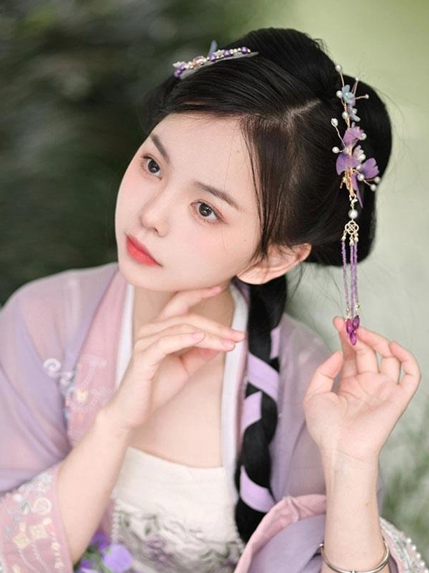Oblique front portrait angle of model in light purple hanfu with ancient braided hair wearing long tassel purple floral and pearl hair design showing graceful movement and delicate cascade of ornament