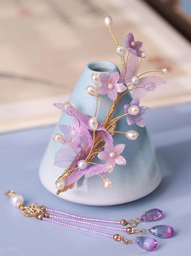 Side-mounted purple floral lolita hair accessory featuring sheer blooms gold metal framework lustrous pearls and multi-strand purple crystal tassel photographed from front angle against gradient blue vase background