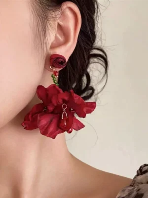 Red fabric flower drop earrings with burgundy rose stud and dark red layered flower pendant featuring green bead accents on model wearing solid off-white background close-up left ear shot gothic lolita elegant jewelry