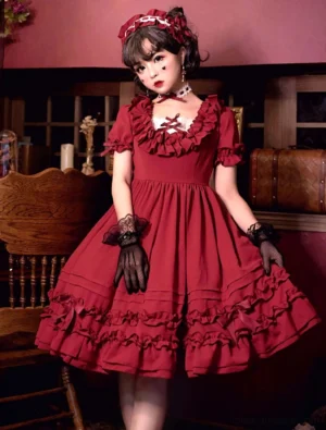 Red sweet lolita one-piece dress with lace-up neckline and tiered ruffled trims, modeled by brunette in classic lolita coordinate with matching hair accessory and mesh gloves, full standing front view