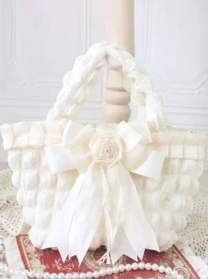 Off-white rococo sweet lolita handbag with cream fabric rose and structured bow detail, quilted textured surface, front view on vintage lace decorated table with pearls