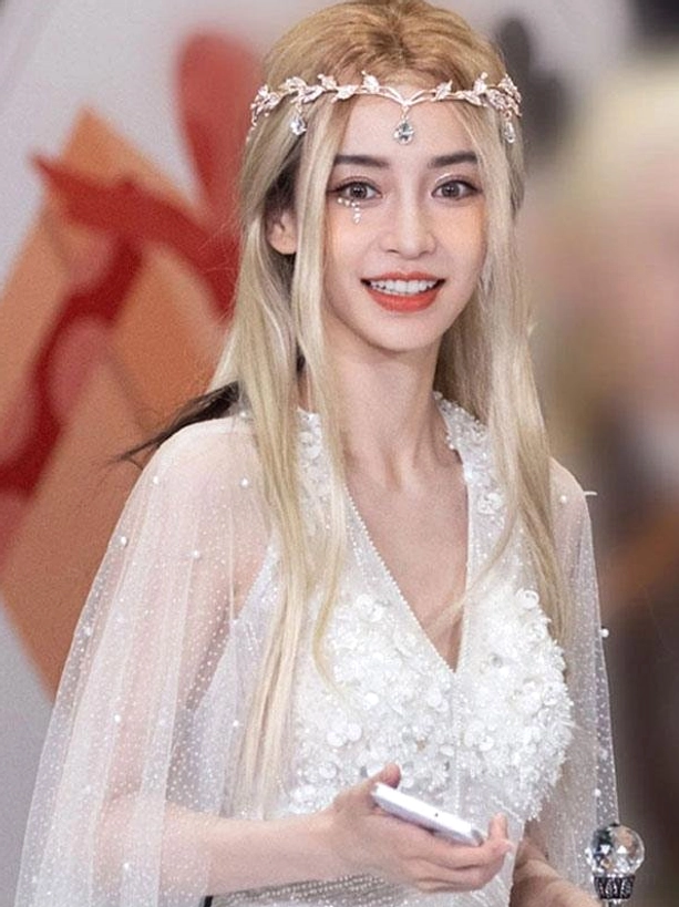 Front three-quarter portrait of light gold rococo lolita wig with long straight blonde hair and crystal leaf crown accessory, model wearing wig against red flag background