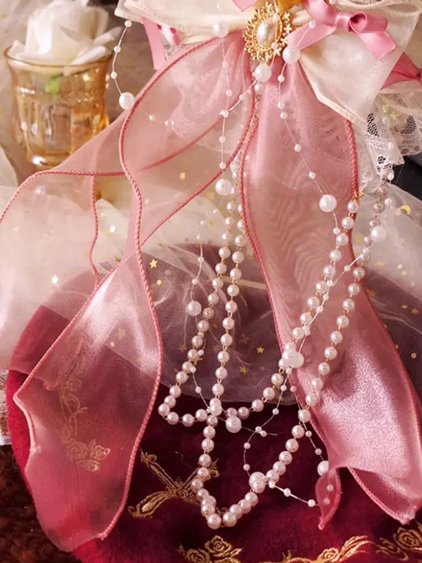 Detailed close-up of romantic pink lolita hair accessory adorned with sheer pink ribbons, delicate pearl chains, small decorative bow and golden brooch detail, showcasing fine craftsmanship on vintage velvet tabletop setting