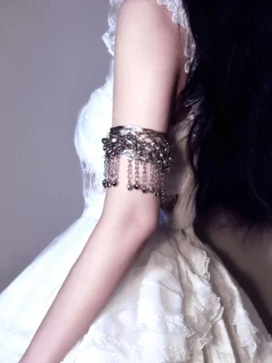 Silver filigree arm cuff back view on upper arm, featuring hanging bell tassels and cross-woven wire hollow frame, worn with white frilly lolita dress, back close-up perspective