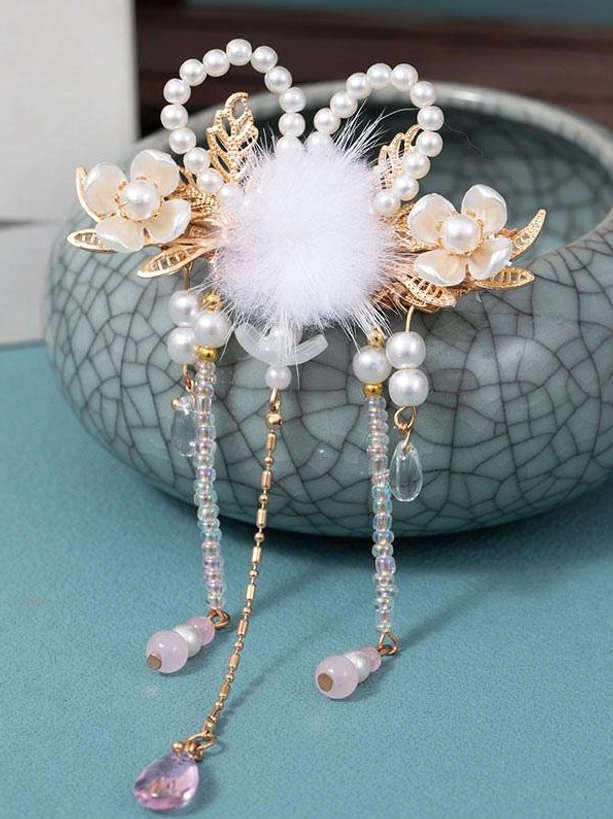 Single decorative hair clip featuring gold metal leaf ornaments, white pom-pom center, beaded hanging tassels, displayed on ceramic vessel, overhead side angle