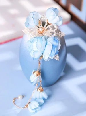 Front oblique view of blue flower tassel hair ornament featuring pearl-inlaid gold center and flowing chain tassel pendants, styled with ceramic vase backdrop