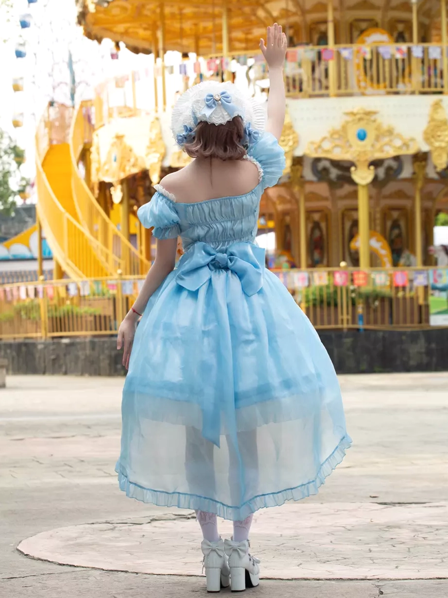 Full back view of sky blue lolita dress with matching bonnet, prominent large back bow, layered ruffled skirt construction, and asymmetrical hemline in amusement park setting