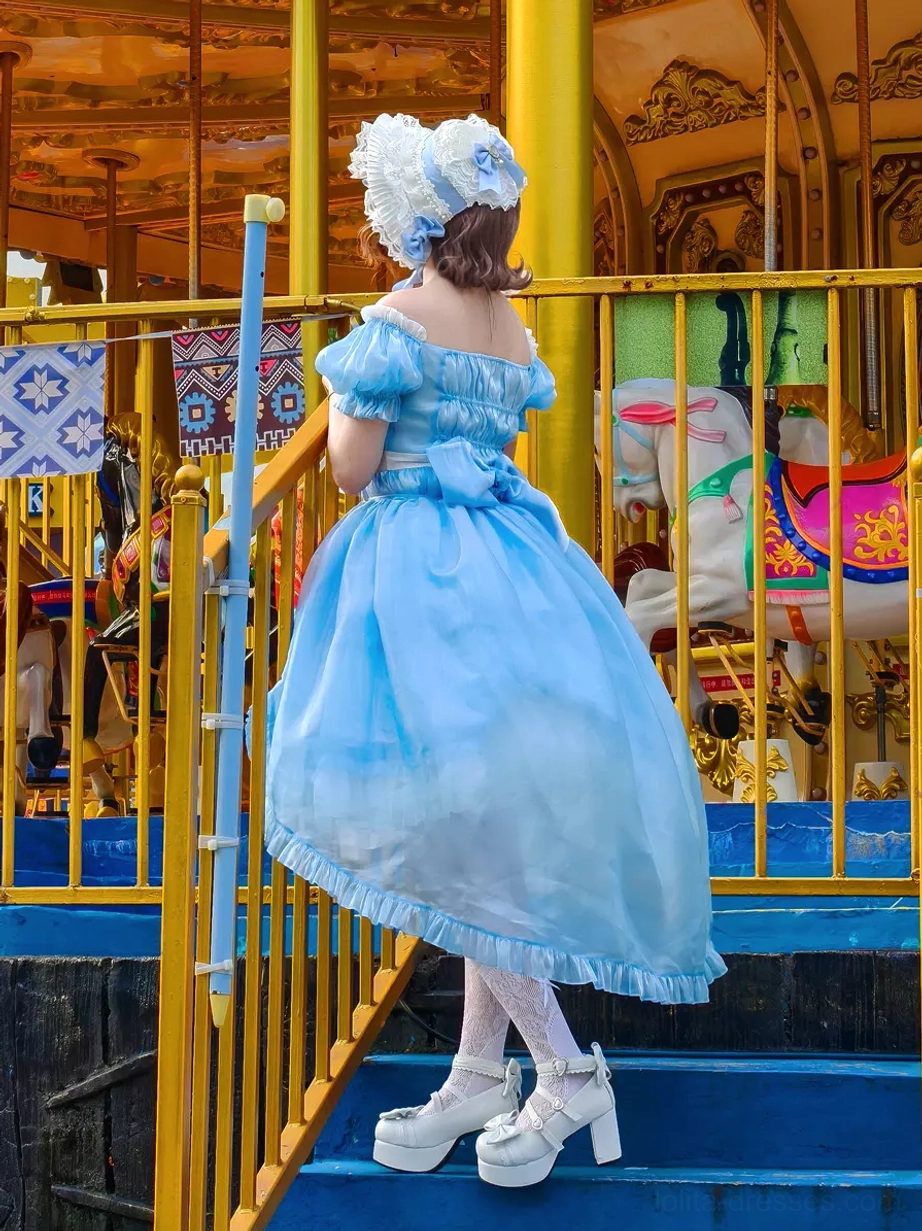Back side view of sky blue lolita dress displaying high-low hemline construction, large decorative back bow, and layered ruffled details in amusement park stairway