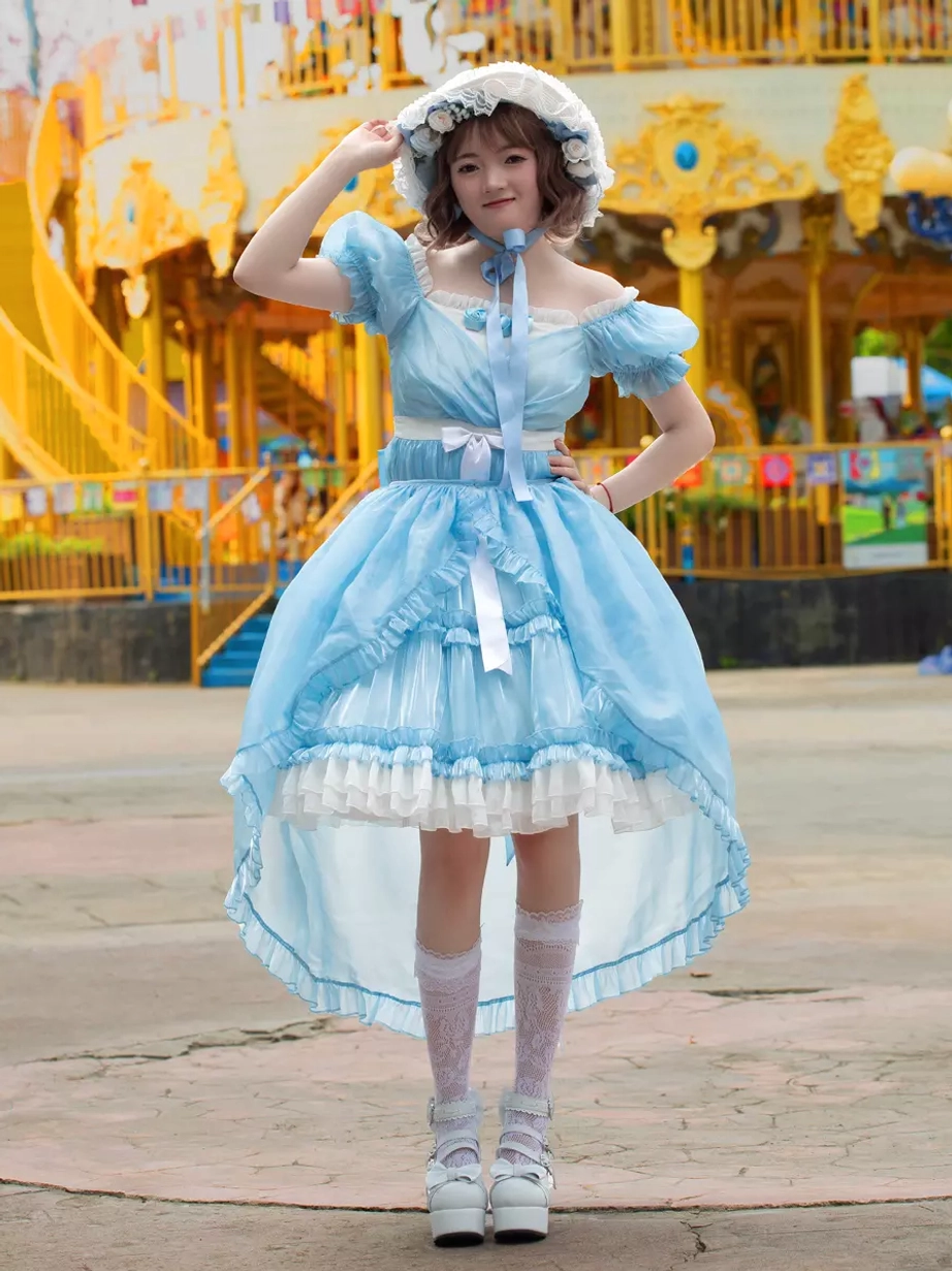 Full front view of sky blue sweet lolita dress with matching floral bonnet, high-low ruffled skirt trim, and layered petticoat effect in amusement park setting