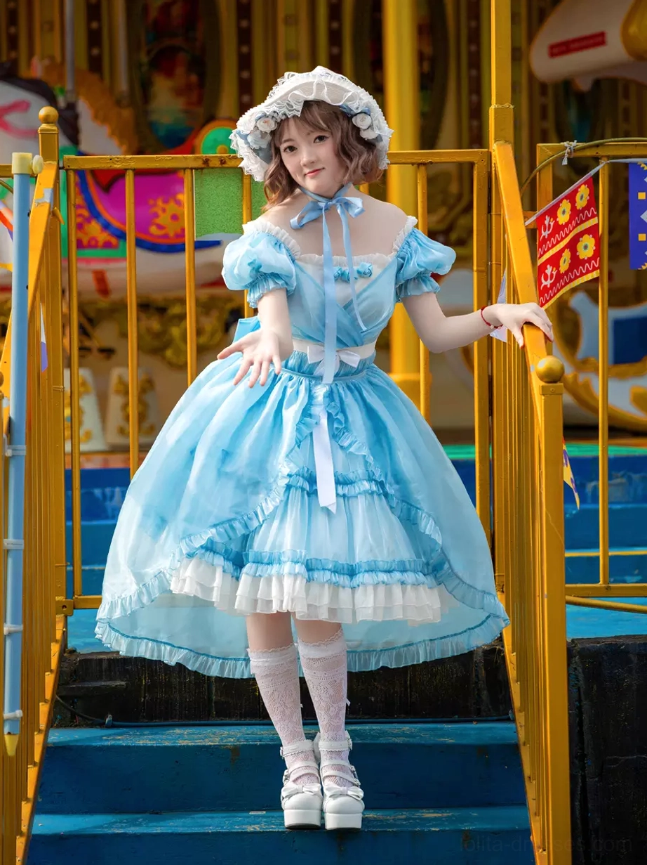 Sky blue sweet lolita dress with off-shoulder puff sleeves and high-low ruffled hem, front three-quarter view showing layered skirt design and satin ribbon accents at amusement park location