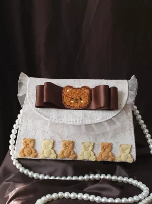 Sweet Lolita bear handbag front flat lay with ruffled lace flap, brown satin bow, pearl chain, and teddy bear trim embellishments on dark brown background