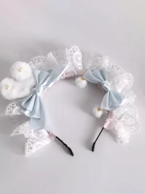 Flat lay side view of pastel blue lolita bunny ear headdress showing delicate white lace ruffles, matching blue satin bows, fluffy pom-pom ears, and hanging pom-pom charms with gold details on light gray background