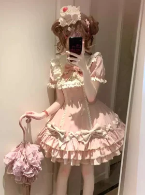 Sweet lolita cake dress with tiered ruffles and short sleeves in soft pink, modeled with matching hat, twin buns hairstyle, and lace parasol accessory - front mirror view