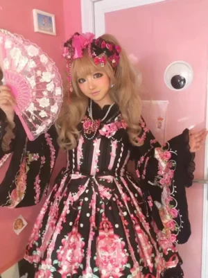 Sweet lolita dress front medium shot featuring black base with pink floral prints, lace collar, long flare sleeves, and lolita girl holding decorative pink folding fan in vintage indoor scene
