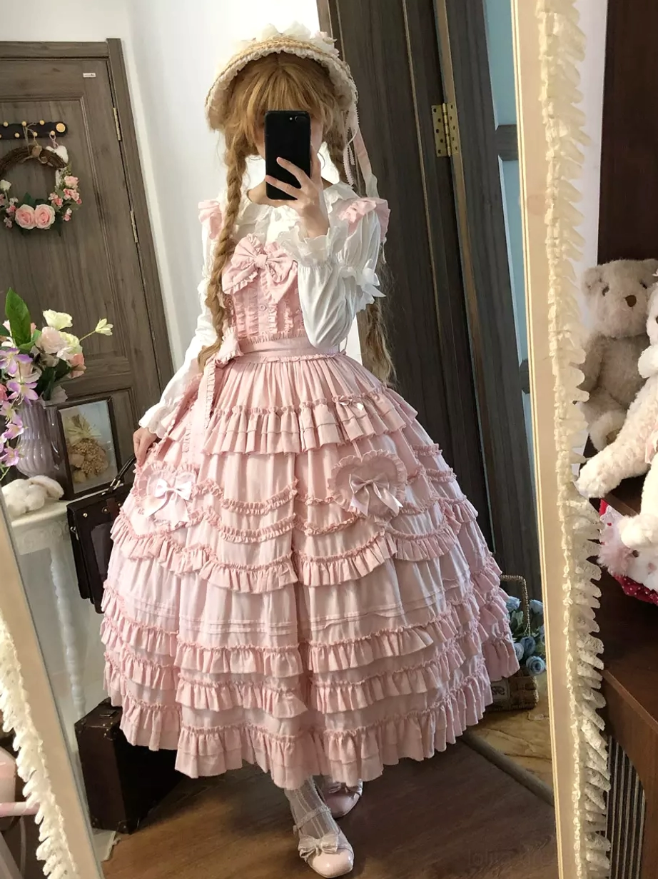 Pink sweet lolita sleeveless dress with tiered ruffles and prominent front bow worn by blonde model wearing straw hat in home entryway