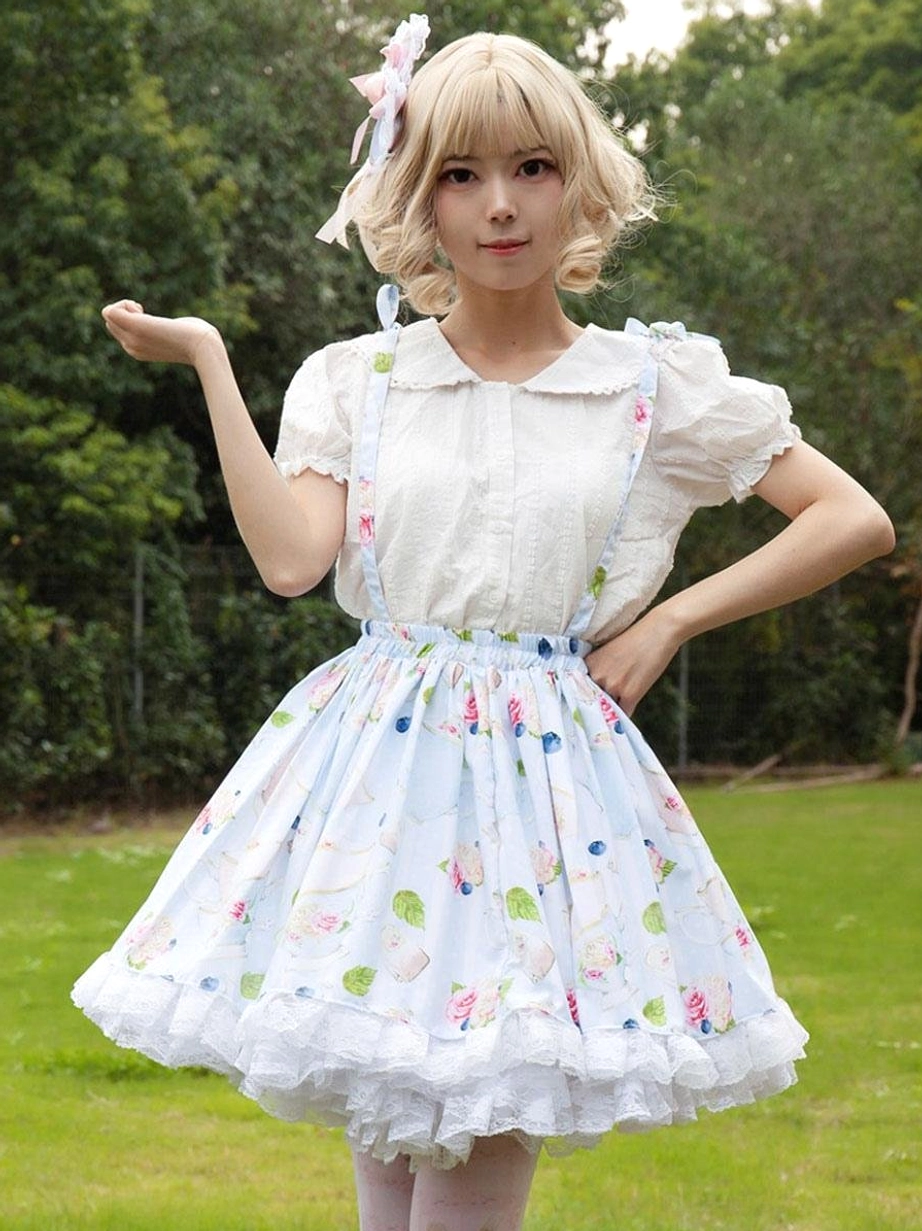 Sweet lolita floral suspender skirt ruffle hem with white puff sleeve blouse casual pose outdoors on lawn blonde model with bow