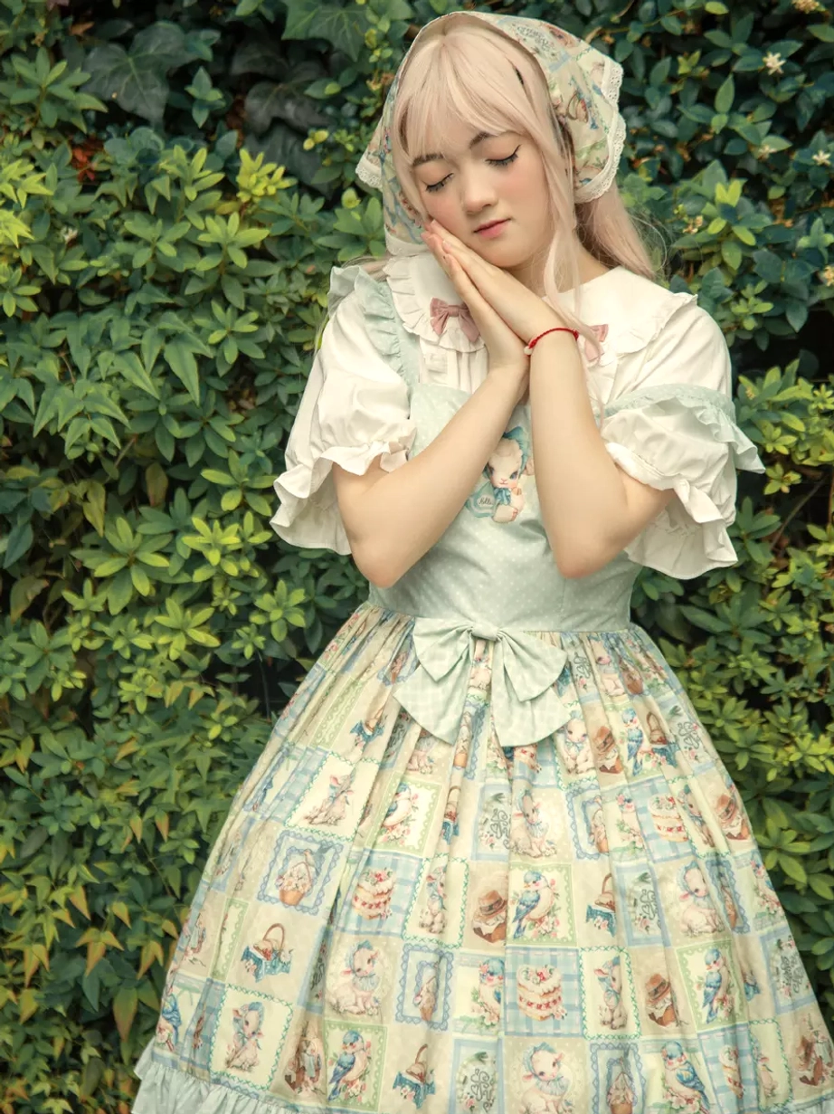 Front eye-level sweet lolita JSK dress set with matching headscarf, puff sleeve blouse, hands-on-cheek pose, and waist bow against green bush backdrop