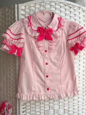 Sweet pink lolita short sleeve blouse with ruffled collar and red bow trims displayed in flat lay on rattan background, showing heart-shaped buttons and detailed frill cuffs