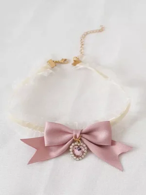 Sweet Lolita pink bow choker with heart-shaped rhinestone gem pendant, off-white ruffled lace strap, dusty pink satin bow, and adjustable gold chain clasp, flat lay front view on white background