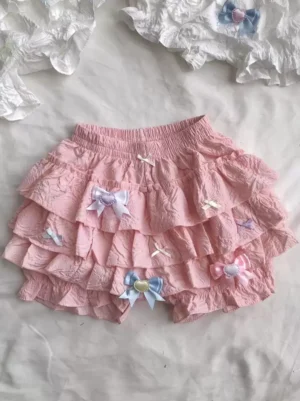 Sweet pink ruffled lolita bloomer shorts flat lay with matching frilled accessories on white bedding showing multi-layer flared ruffles and textured crepe fabric