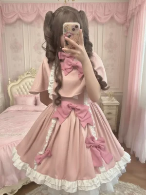 Sweet pink lolita dress with detachable cape front view featuring multiple large pink fabric bows, white ruffled collar with pink trim, and flared skirt silhouette on model in vintage pink bedroom setting