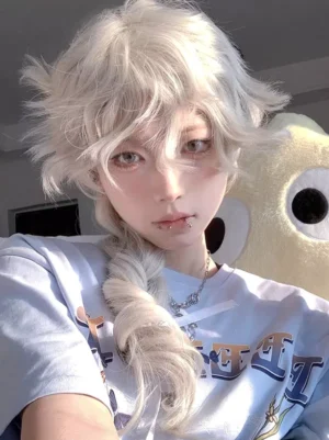Front close-up selfie of tousled light gold Ouji Lolita wig with messy layered styling and long braid, worn with light blue printed shirt against yellow plush toy background