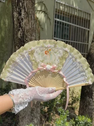 Vintage oil painting lady print lace ruffle folding hand fan with pink floral bow, held by white lace-gloved hand in outdoor garden setting with trees and building backdrop, featuring green ruffled lace border and romantic classical portrait design perfect for lolita fashion
