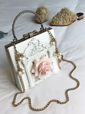 White gothic lolita box handbag with carved frame, pink rose and pearl strap, flat lay styling with fuzzy slippers props on neutral background